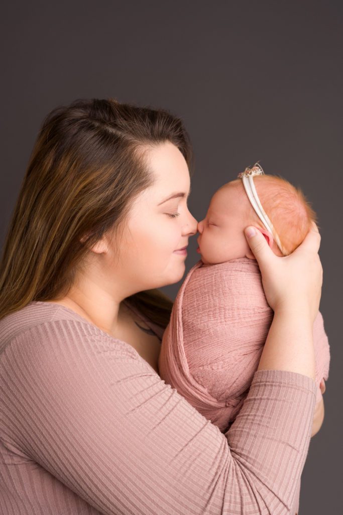 Studio newborn photo of little baby girl in blush pink wrapped up, mother daughter photo touching noses with thunder gray backdrop