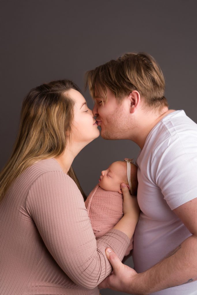 Studio newborn photo of little baby girl in blush pink wrapped up, parent mom and dad photo with thunder gray backdrop