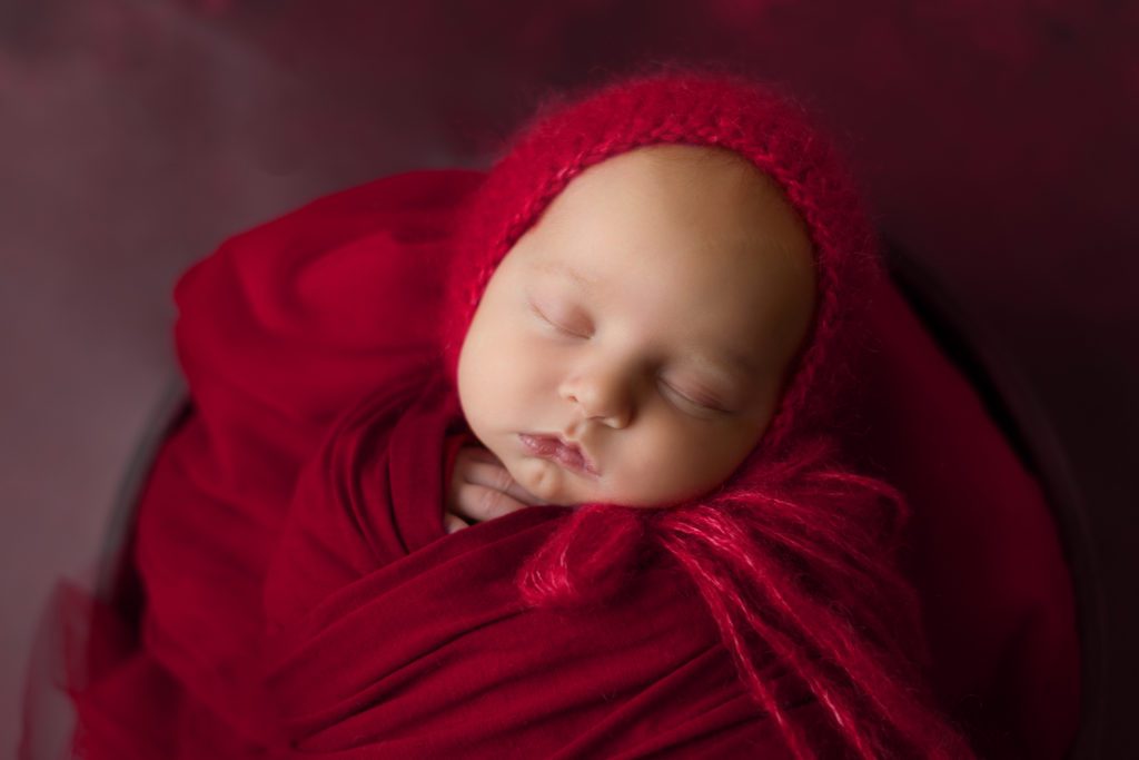 Studio newborn photo of little baby girl with red bonnet and wrap in a basket of flowers