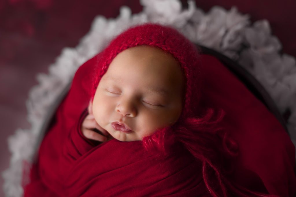 Studio newborn photo of little baby girl with red bonnet and wrap in a basket of flowers